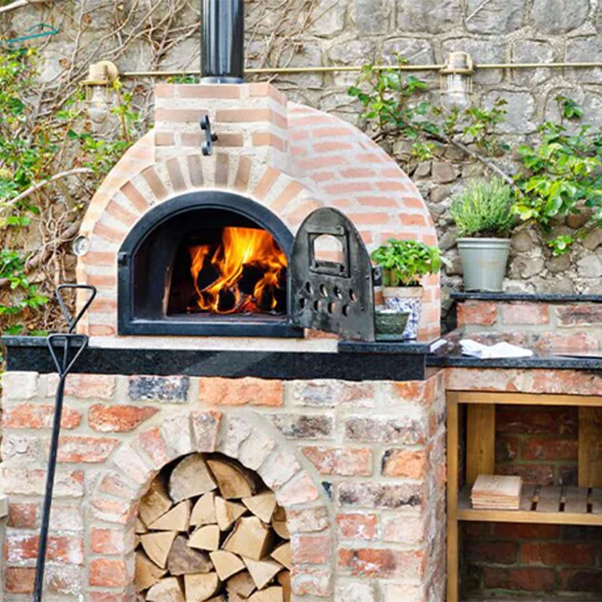 Brick outdoor oven with fire inside, surrounded by stone wall and plants