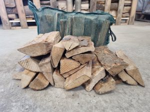 Stack of firewood in front of a green basket on a concrete floor with wooden pallets in the background.